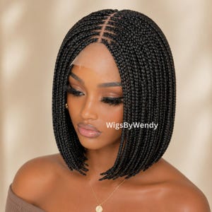 Puede incluir: Una peluca negra tipo bob trenzada con raya al medio y base de encaje frontal. El cabello está peinado en microtrenzas uniformes y finas que llegan hasta la barbilla. El texto WigsByWendy es visible en el lado derecho de la imagen.