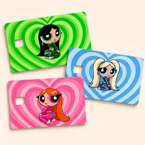 May include: Three rectangular credit card skins featuring cartoon characters in matching tracksuits with the word Juicy printed on the back. Each card has a heart-patterned background in green, blue, or pink, and a small square cutout on the left side.