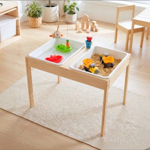 May include: A light-colored wooden sensory table featuring two removable white plastic bins. One bin contains water with a green toy boat, a red toy boat, and a fishing rod. The other bin contains sand with two yellow and black toy dump trucks.