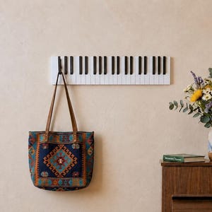 May include: A wall-mounted coat rack designed as a horizontal piano keyboard with alternating black and white keys. The rack features a single dark hook holding a rectangular tote bag with a blue, orange, and brown geometric pattern.