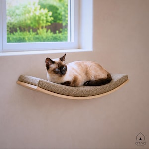 Peut inclure: Un perchoir pour chat mural et incurvé, doté d'un cadre en bois clair et d'un coussin en tissu beige texturé. Un chat siamois aux extrémités brun foncé et aux yeux bleus est allongé sur le perchoir, situé sous une fenêtre.
