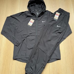 May include: A black Nike tracksuit consisting of a hooded zip-up jacket and matching athletic pants. Both pieces feature a white Nike swoosh logo and attached retail tags. The jacket has a full-length zipper and a hood, while the pants have elastic cuffs.