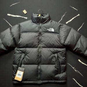 Op de afbeelding: Een zwarte gewatteerde jas met een hoge kraag en een ritssluiting over de gehele lengte. Het The North Face logo is in het wit geborduurd op de linkerborst. De jas heeft horizontale gewatteerde panelen en een bruin kartonnen productlabel.