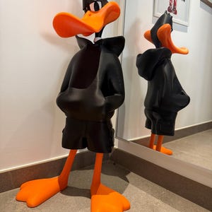 Puede incluir: Una figura de pie del personaje de dibujos animados Pato Lucas, que lleva una sudadera con capucha negra y pantalones cortos negros. El personaje tiene un pico naranja brillante y grandes pies palmeados de color naranja. La figura tiene un acabado mate liso con una superficie reflectante visible en el espejo del fondo, capturando la postura icónica y los rasgos faciales distintivos del personaje.
