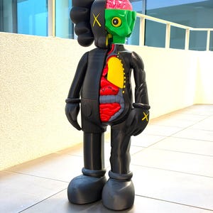 Può includere: Una figura d'arte da collezione nera con un design sezionato. Un lato mostra un esterno nero solido con segni a X gialli sugli occhi e sulle mani, mentre l'altro lato rivela una testa verde con un cervello rosa e organi interni in rosso, giallo e grigio.
