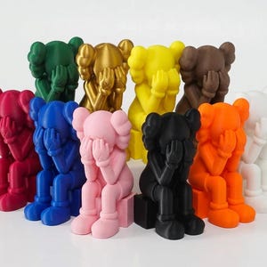 Può includere: Una collezione di undici statuette in stile KAWS in vari colori solidi, tra cui rosso, verde, oro, giallo, marrone, blu, rosa, nero, arancione e bianco. Ogni figura è raffigurata in posizione seduta con le mani che coprono il viso.