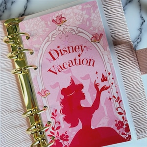 Peut inclure: Un agenda rose avec un mécanisme de reliure à six anneaux dorés. La couverture présente une silhouette de personnage aux longs cheveux portant une couronne, entourée d'oiseaux, de fleurs et d'un château. Le texte Disney Vacation est en rouge.