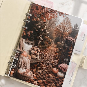 Può includere: Un'agenda ad anelli color crema con una pagina divisoria che mostra una scena di giardino autunnale. L'immagine presenta una panchina in legno, zucche e foglie cadute. Sul lato destro sono visibili due divisori con le etichette agosto e settembre.