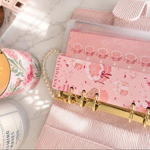 Puede incluir: Una carpeta de anillas de color rosa claro con un mecanismo de metal dorado y un inserto rosa con el texto BIRTHDAYS e ilustraciones de pasteles y regalos. Al lado hay una vela encendida en un frasco de vidrio floral y una segunda vela.
