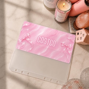 Op de afbeelding: Een doorzichtige plastic ordnerhoes met een roze marmerontwerp, twee lichtroze strikken aan de zijkanten en het woord COSTCO in witte hoofdletters in het midden. De hoes heeft een reeks ronde gaten langs de onderste rand.