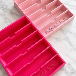 May include: Two rectangular cash tray organisers with four horizontal compartments each. One tray is light pink and the other is hot pink. Both trays feature the text Budget Babe Studio embossed on the bottom right corner of the front compartment.