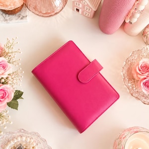 Puede incluir: Una agenda personal de color rosa intenso vibrante con una cubierta lisa de color sólido y un cierre de correa rectangular a juego. La agenda tiene forma rectangular y presenta un diseño limpio sin patrones, texto ni logotipos visibles en el exterior.