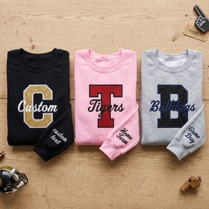 Peut inclure: Trois sweat-shirts à col rond en noir, rose clair et gris chiné. Chacun présente une grande lettre universitaire scintillante avec un texte en écriture brodé au centre et du texte personnalisé brodé sur le poignet.