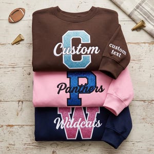 May include: Three crewneck sweatshirts in brown, pink, and navy blue. Each features a large glittery letter and script text: brown with light blue 'Custom', pink with blue 'Panthers', and navy with pink 'Wildcats'. Sleeve has 'custom text'.
