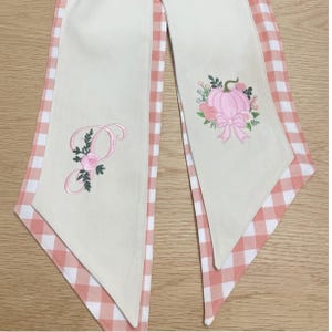 Pu&ograve; includere: Due lunghe strisce di tessuto con un pannello centrale color crema e un bordo a quadretti vichy color pesca e bianco. La striscia sinistra presenta una lettera P ricamata con rose rosa e foglie verdi. La destra mostra una zucca rosa.