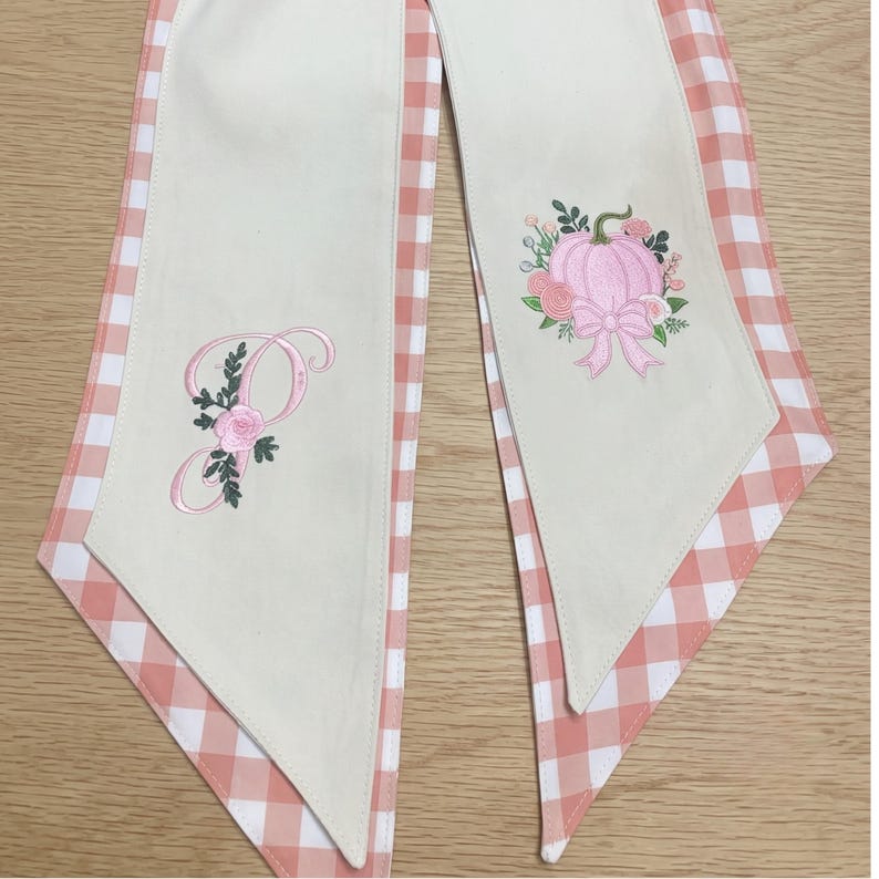 Pu&ograve; includere: Due lunghe strisce di tessuto con un pannello centrale color crema e un bordo a quadretti vichy color pesca e bianco. La striscia sinistra presenta una lettera P ricamata con rose rosa e foglie verdi. La destra mostra una zucca rosa.