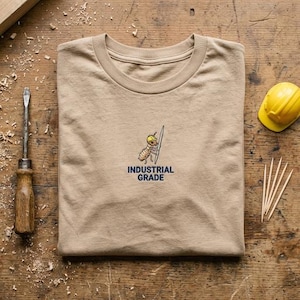 May include: A tan crew neck t-shirt featuring an embroidered graphic of an ant wearing a yellow hard hat and holding a tool, with the text "INDUSTRIAL GRADE" in dark blue lettering centered below the illustration on the chest area.