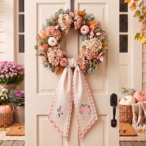 Pu&ograve; includere: Una ghirlanda circolare con zucche artificiali rosa e bianche, fiori di ortensia essiccati e foglie autunnali. Un lungo nastro in tessuto color crema con bordo a quadretti vichy rosa pende dal basso, con una lettera P e una zucca ricamate.