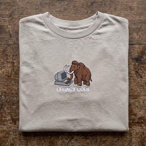 May include: A beige crew neck t-shirt featuring an embroidered graphic of a brown woolly mammoth standing next to a vintage grey computer. Below the graphic, the text "LEGACY CODE" is embroidered in white capital letters on the chest.