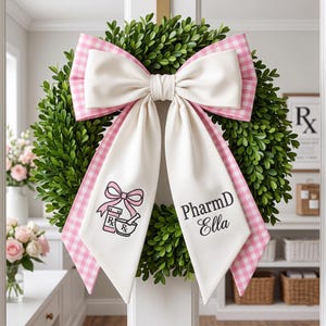 May include: A green boxwood wreath adorned with a large layered bow featuring white fabric and pink gingham ribbon. The white ribbon tails display a graphic of a pink bow, a prescription bottle, and a mortar and pestle, with the text PharmD Ella.