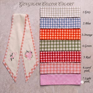 Pu&ograve; includere: Una tabella colori Gingham con una lunga striscia di tessuto color crema con bordi a quadretti arancioni, ricamata con una lettera P e una zucca. A destra ci sono sette campioni di tessuto in grigio, blu, arancione, verde, rosso, rosa carol e rosa chiaro.