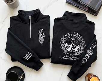 May include: Two black quarter-zip sweatshirts featuring white graphic prints. One shows a hooded figure holding a mug and text on the sleeve. The other displays a mountain scene with the text Velaris City of Starlight and moon phases on the sleeve.