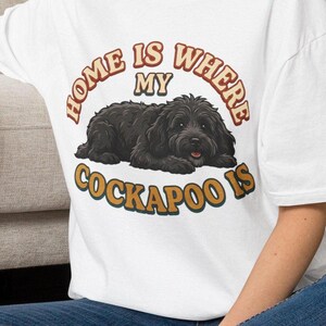 Könnte beinhalten: Ein weißes T-Shirt mit einer Grafik eines liegenden schwarzen Cockapoo-Hundes. Der Text "HOME IS WHERE MY COCKAPOO IS" ist in einer geschwungenen Retro-Schriftart über und unter der Hundeillustration in Orange-, Braun- und Petroltönen gedruckt.