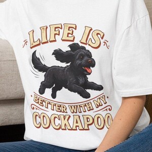 May include: A white t-shirt featuring a graphic of a black Cockapoo dog running with its tongue out. The text above the dog reads LIFE IS and the text below the dog reads BETTER WITH MY COCKAPOO in a stylised, vintage-inspired brown and tan font.