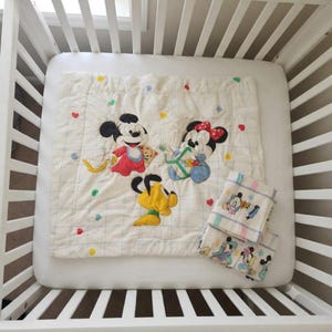 May include: A white quilted baby blanket featuring embroidered images of Mickey Mouse, Minnie Mouse, and Pluto, accented with a grid pattern and small colorful hearts and dots. A smaller matching folded blanket is placed on the side.
