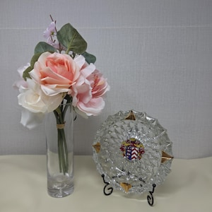 May include: A clear glass cylindrical vase containing a bouquet of artificial pink and white roses with green leaves, positioned next to a circular, faceted clear glass ashtray featuring a colorful heraldic crest and gold-toned accents.