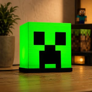 Könnte beinhalten: Eine quadratische, leuchtend grüne Lampe mit dem ikonischen schwarzen, pixeligen Gesicht eines Creepers aus Minecraft. Die Lampe hat einen soliden schwarzen Sockel und strahlt ein helles, gleichmäßiges Licht über die Vorderseite aus.