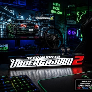 May include: A rectangular LED sign displaying the Need for Speed Underground 2 logo with white lettering and a red number 2. The sign is positioned on a desk in front of a gaming setup featuring a steering wheel controller and a computer monitor.