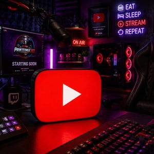 May include: A rectangular light-up sign featuring the iconic red YouTube logo with a white play button in the centre. The sign is positioned on a desk in a gaming setup, surrounded by computer monitors, a keyboard, and neon wall signage.