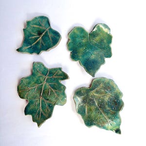 May include: Four ceramic dishes shaped like leaves, each featuring a glossy, mottled green glaze with visible dark veining and textured surfaces. The leaf-shaped plates vary slightly in form and size, showcasing organic edges and detailed vein patterns.