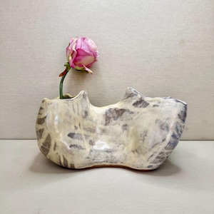 May include: A low-profile, elongated ceramic vase with an irregular, organic shape and a cream-coloured glaze featuring dark grey, mottled, and streaked patterns. A single pink rose is placed in the opening on the left side of the vessel.