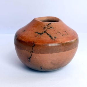 May include: A small, round, terracotta-coloured clay pot with a wide, tapered opening. The surface features a smooth, matte finish with a horizontal band of darker brown clay and distinct, thin, black, branching crack-like markings.