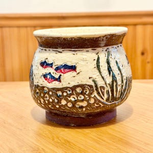 Op de afbeelding: Een ronde keramische pot met een brede rand, voorzien van een getextureerd crèmekleurig bovengedeelte versierd met kleine blauwe en rode vissen en donkergroen zeewier, en een donkerbruin ondergedeelte met een verhoogd, cirkelvormig patroon.