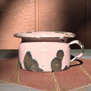 May include: A vintage light pink enamel chamber pot featuring a wide rim and a single side handle. The exterior surface displays several dark, distressed patches of exposed metal and rust, showing significant wear and age on the edges.