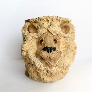 May include: A ceramic mug shaped like a lion head with a textured, cream-colored mane. The lion features a smooth tan face, dark brown eyes, a black nose, and a small tongue protruding from the mouth. The vessel has a cylindrical form.