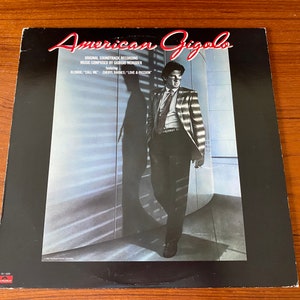 May include: A black vinyl record album cover with a white and red title "American Gigolo". The cover features a man in a striped jacket standing in a room with a large window. The text on the cover reads "Original Soundtrack Recording", "Music Composed by Giorgio Moroder", "Featuring", "Blondie/Call Me", "Cheryl Barnes/Love & Passion".