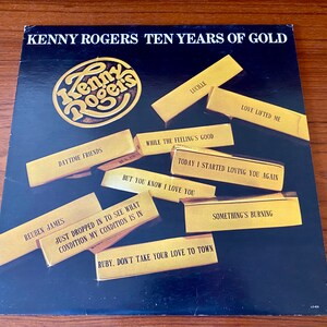 May include: A black vinyl record album cover with gold lettering that reads "Kenny Rogers Ten Years of Gold". The album cover features a gold circular logo with the text "Kenny Rogers" inside. The album cover also features a collage of gold rectangles with song titles printed on them, including "Lucille", "Love Lifted Me", "Daytime Friends", "While the Feeling's Good", "Today I Started Loving You Again", "But You Know I Love You", "Reuben James", "Just Dropped In to See What Condition My Condition Is In", "Ruby. Don't Take Your Love to Town", and "Something's Burning".