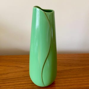 May include: A tall, light green ceramic vase with a curved design on the front. The vase has a smooth, glossy finish and a unique, asymmetrical top. The vase is displayed on a wooden surface.