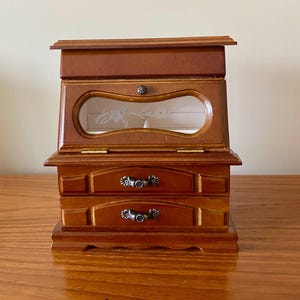 May include: A wooden jewelry box with a warm brown finish, featuring a top compartment with a glass window etched with a floral design, and two lower drawers equipped with decorative silver-toned metal handles. The structure is rectangular.