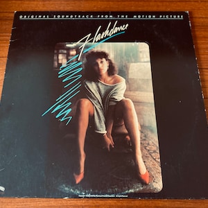 May include: Album cover for the "Flashdance" soundtrack. The cover features a woman in a gray sweater and red heels. The title is in a stylized font with turquoise accents against a black background. The text "ORIGINAL SOUNDTRACK FROM THE MOTION PICTURE" is at the top.