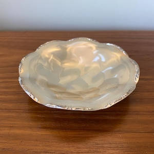 May include: A small, round, shallow dish with a reflective, metallic silver-toned surface. The bowl features a smooth, undulating interior and a decorative, scalloped edge with intricate, fine-line detailing along the entire outer rim.
