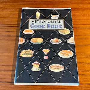 May include: A black and gold cookbook with the title "Metropolitan Cook Book" on the cover. The cover features illustrations of various foods, including a pie, a bowl of soup, a plate of fish, and a plate of pastries.