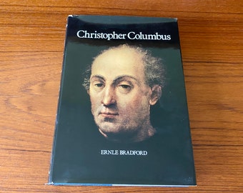 May include: A hardcover book titled "Christopher Columbus" by Ernle Bradford. The book features a portrait of Christopher Columbus on a black background. The book is resting on a wooden surface.