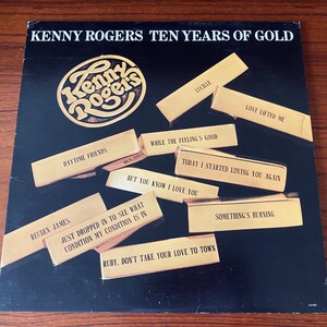May include: A black vinyl record album cover with a gold circle featuring the text "Kenny Rogers" in a gold font. The album title is "Kenny Rogers Ten Years of Gold". The cover features a collage of gold rectangles with song titles printed in black text, including "Lucille", "Love Lifted Me", "Daytime Friends", "While the Feeling's Good", "Today I Started Loving You Again", "But You Know I Love You", "Reuben James", "Just Dropped In to See What Condition My Condition Is In", "Ruby, Don't Take Your Love to Town", and "Something's Burning".