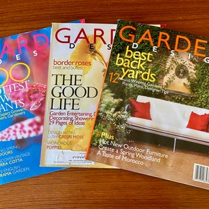 May include: Three magazines with the title "Garden Design" are displayed on a wooden surface. The magazines feature colorful covers with text such as "100 Hottest New Plants" and "Best Back Yards". The magazines are for gardening enthusiasts.