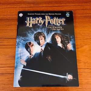 May include: A CD of "Harry Potter and the Chamber of Secrets" soundtrack. The cover features the film's title in gold lettering, with images of Harry, Ron, and Hermione. The CD case is on a wooden surface.